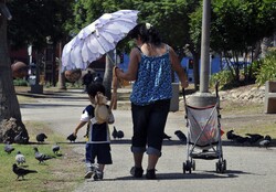 Mother walks along side of her child to shield him from the sun with her umbrella, at a park