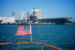 United States flag waves in front of water and a view of a Navy Aircraft Carrier