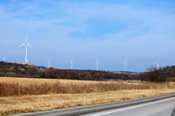 A line of large white wind turbines setting on hill tops along an Oklahoma country road, with a blue sky background.