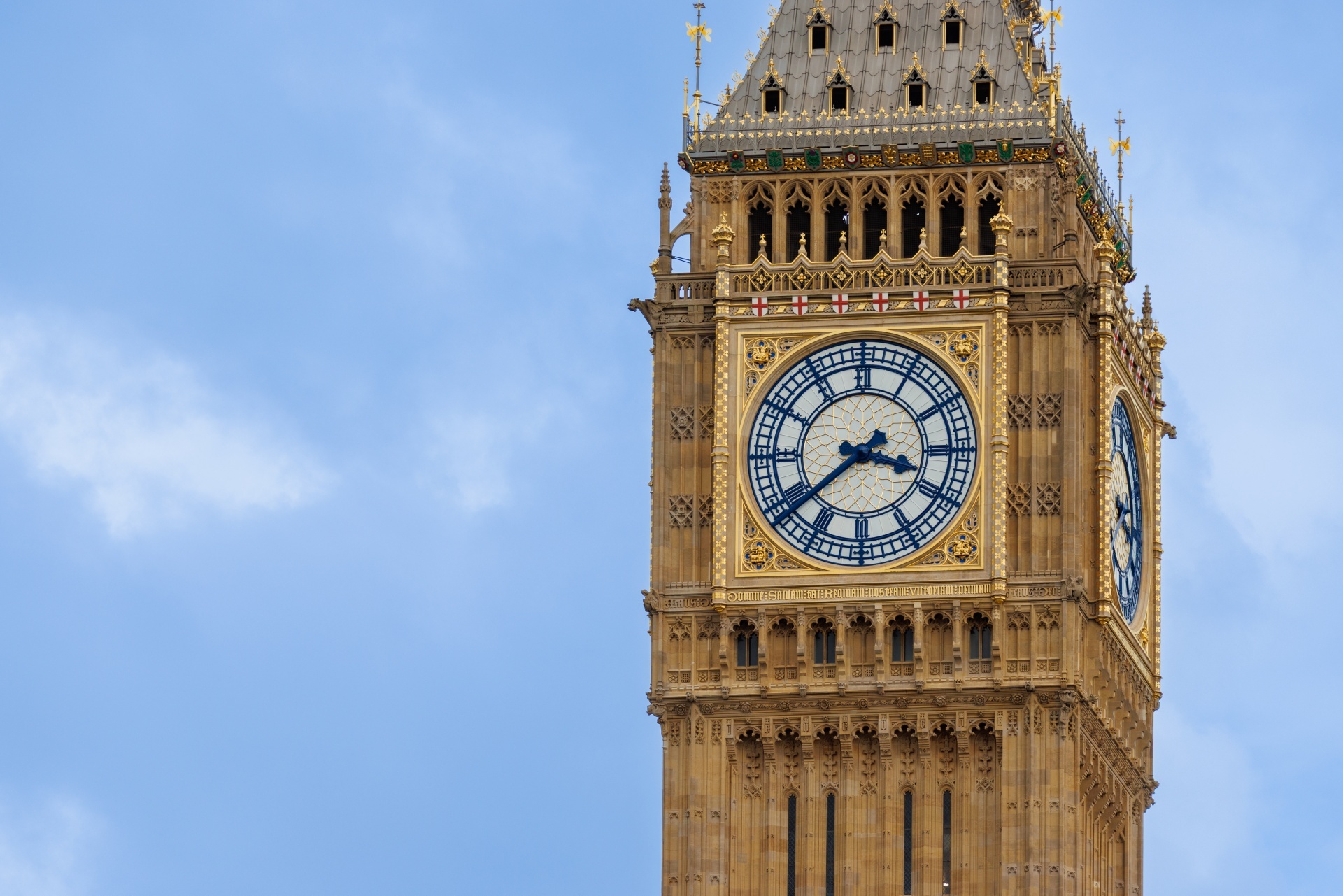 A close-up view of a golden-trimmed Big Ben clock tower in London with a large white clock face set against a clear blue sky.