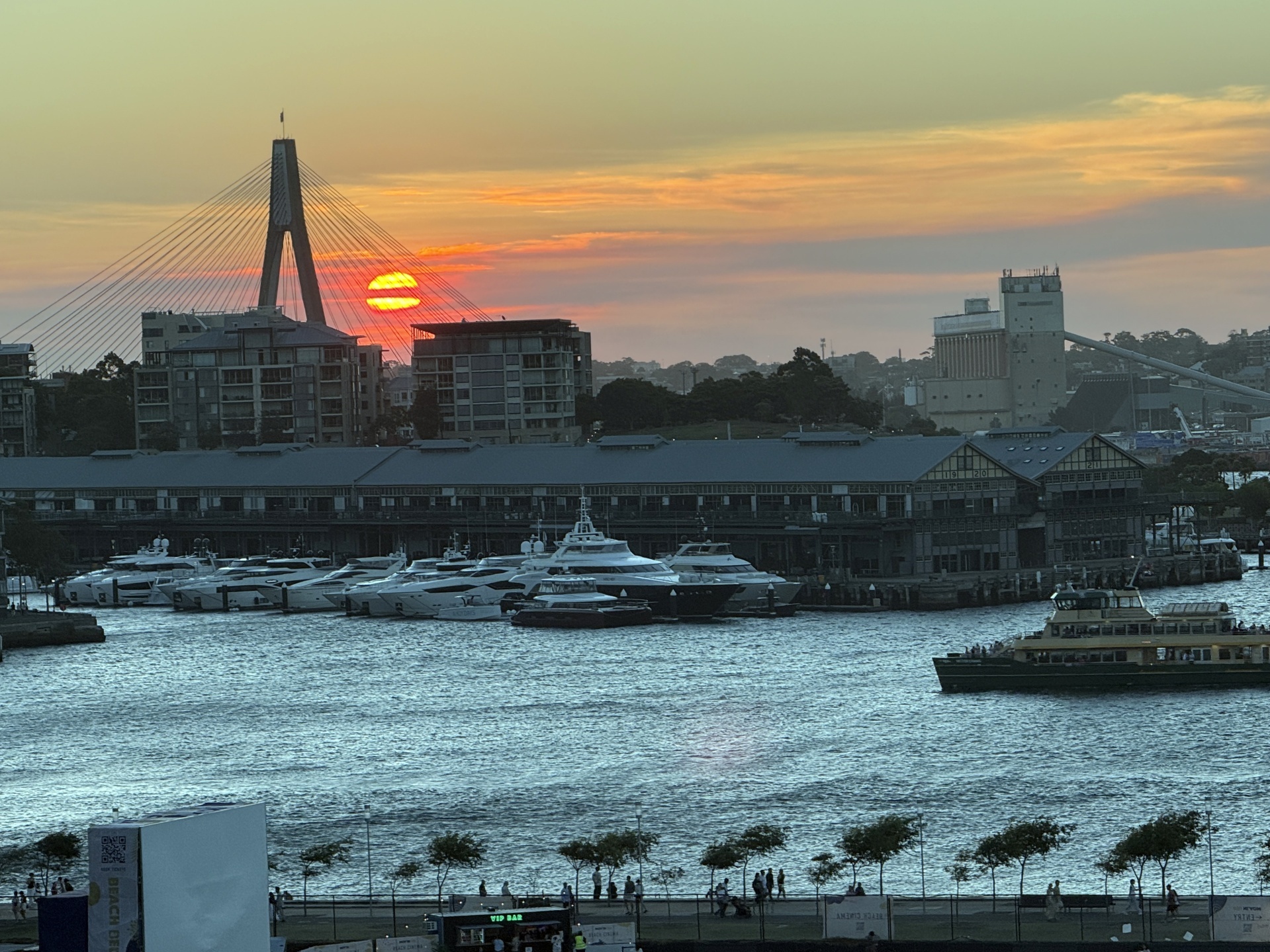 Sydney cityscape showcasing Anzac Bridge, harbor, and boats at sunset. Waterfront buildings with a ferry moving on the water