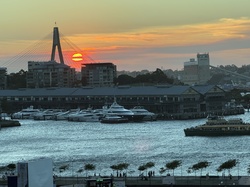 Sydney cityscape showcasing Anzac Bridge, harbor, and boats at sunset. Waterfront buildings with a ferry moving on the water