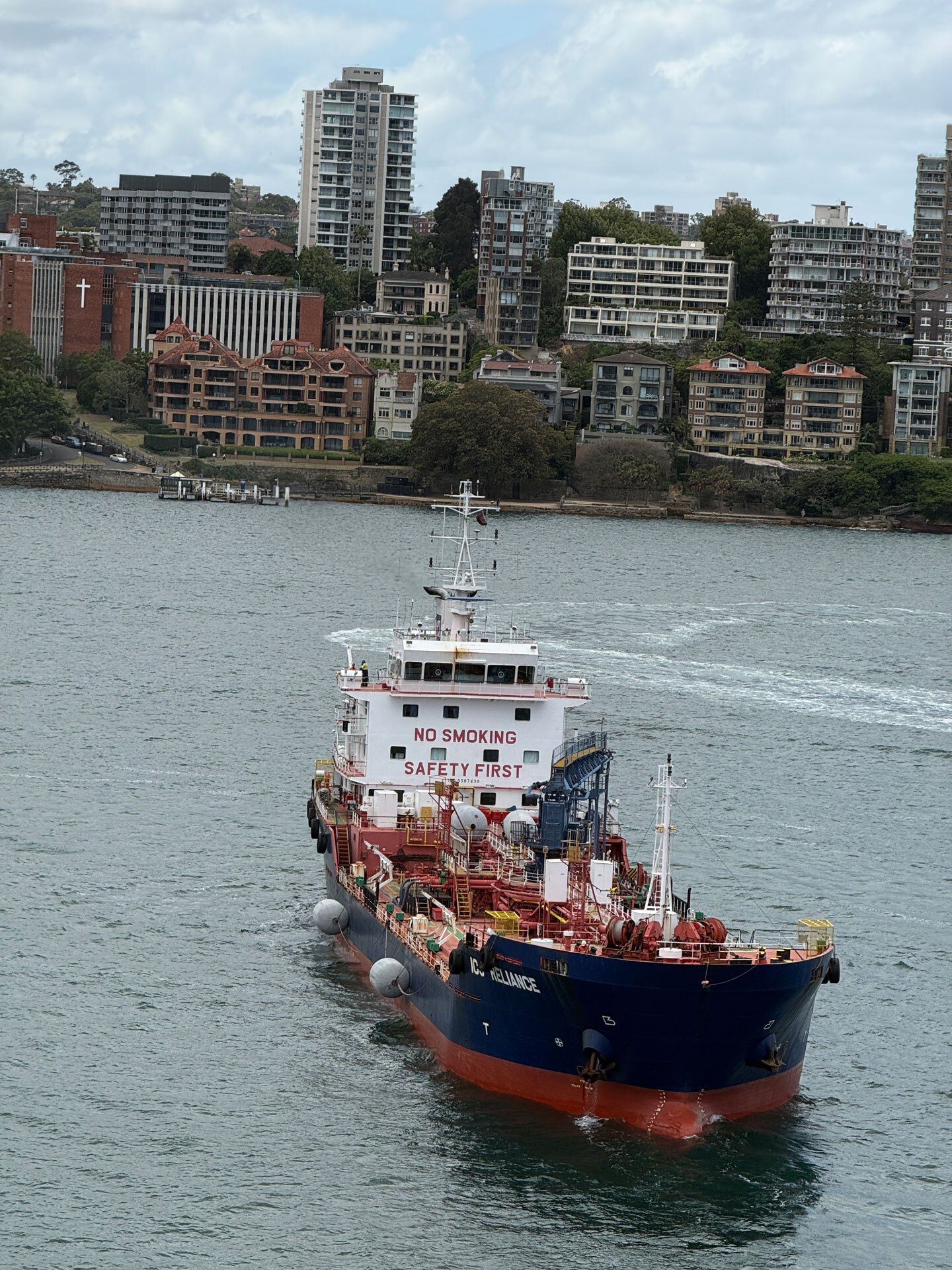 Tanker ship traveling through Sydney Harbour water with city buildings lining the shore as viewed from a departing cruise ship balcony