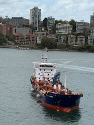 Tanker ship traveling through Sydney Harbour water with city buildings lining the shore as viewed from a departing cruise ship balcony