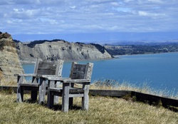 Two rustic wooden chairs facing coastal cliffs and the calm ocean, representing relaxation, vista, and travel destination