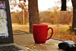 A coffee cup sitting beside a lap top computer, on an outdoor table, with autumn trees in background.