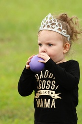 A cute little girl with a princess crown on, is blowing up a purple balloon, on a green blurred background.