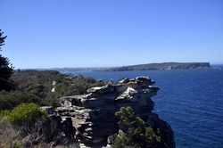 Sandstone cliffs overlook the vast blue ocean and distant Sydney Harbour, offering a wide coastal panorama on a clear day